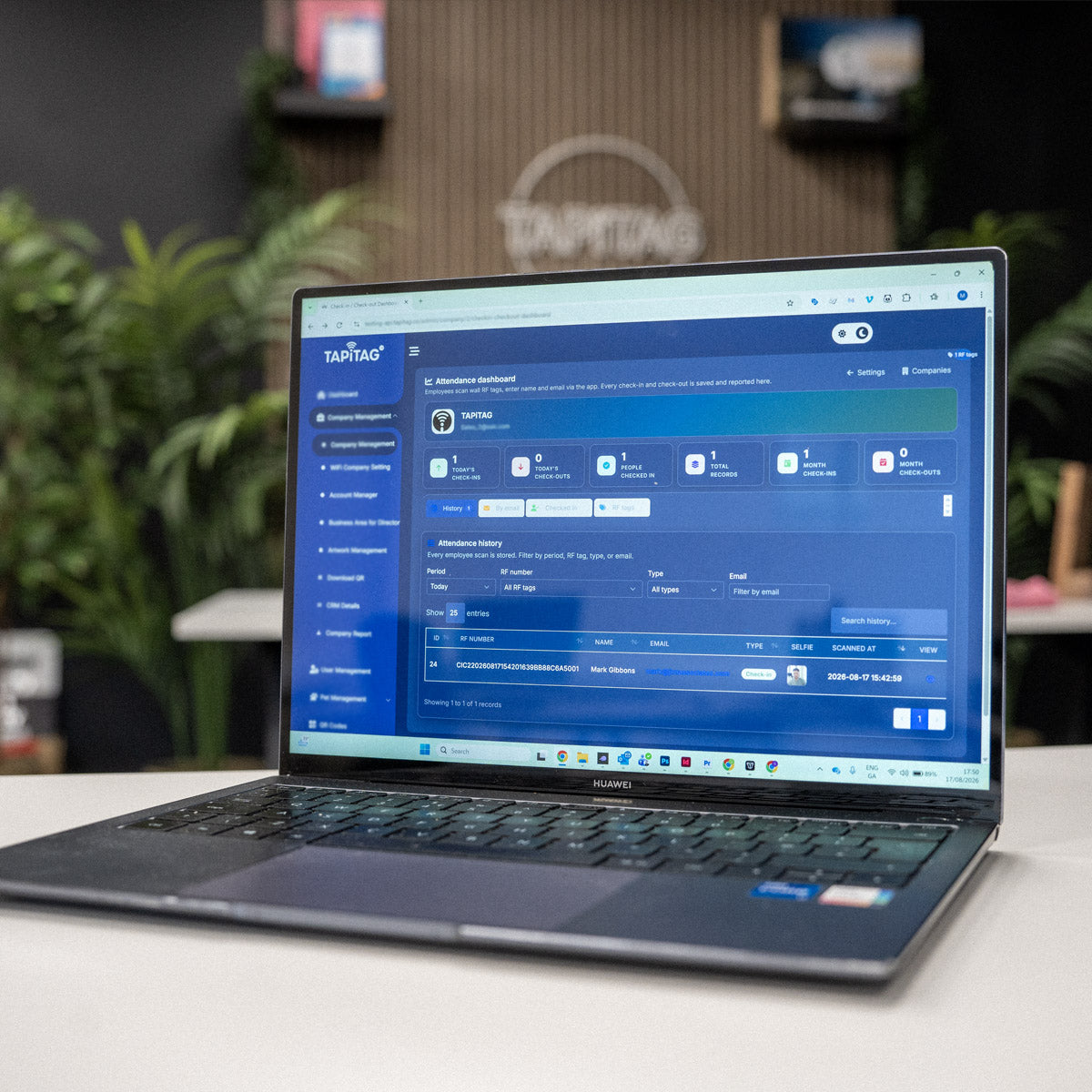 A dark gray HP laptop open on a light-colored table, displaying a blue software dashboard on its screen. The background is a softly blurred office environment with green plants and a wooden wall.