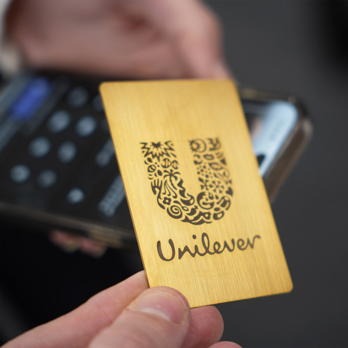 Close-up of a premium Gold TAPiTAG metal digital business card wth unileaver logo tapping a smartphone for instant contact sharing NFC