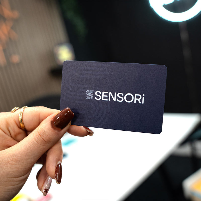 Hand holding a TAPiTAG Digital Business card  with 'SENSORi' branding against a blurred indoor background