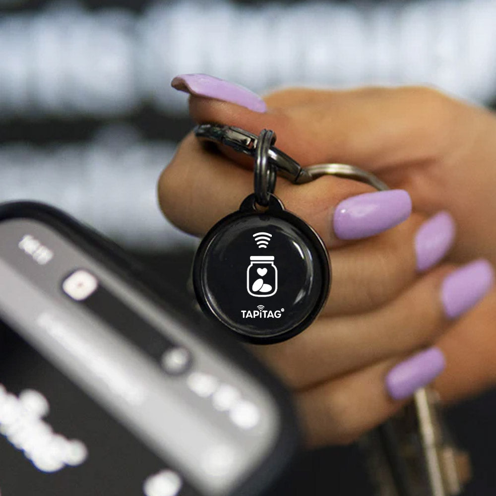 Hand holding a keychain with a Tapitag device in front of a smartphone.