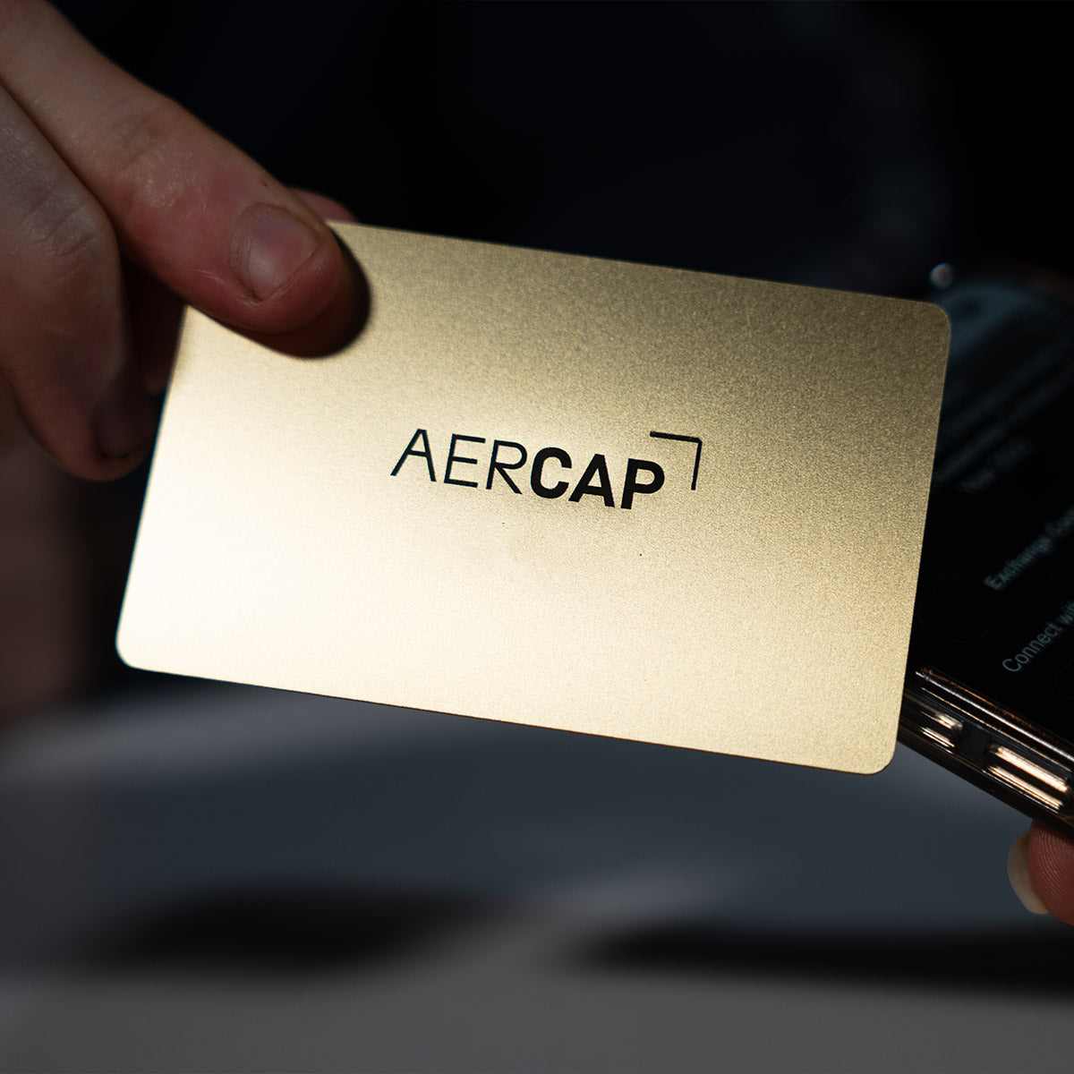 Close-up of a premium Gold TAPiTAG metal digital business card wth aercaplogo tapping a smartphone for instant contact sharing NFC