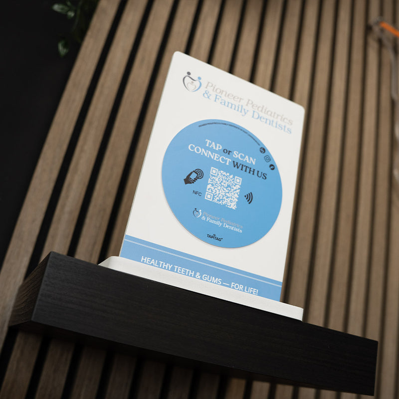 QR code sign on a stand with a wooden background. Promotional sign for Pioneer Pediatrics & Family Dentists with a QR code on a blurred background. TAPiTAG NFC TAG Display stand