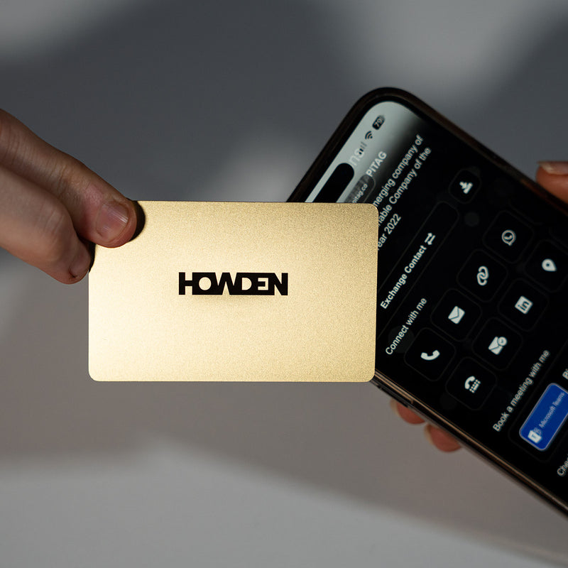 Close-up of a premium Gold TAPiTAG metal digital business card wth Howden logo tapping a smartphone for instant contact sharing NFC