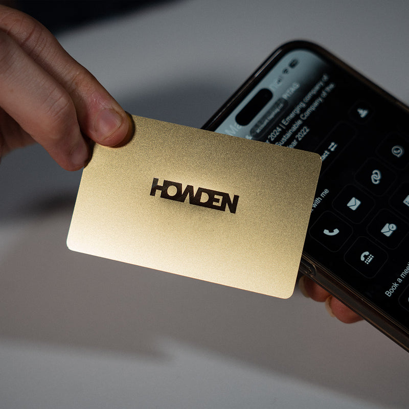 Close-up of a premium Gold TAPiTAG metal digital business card wth Howden logo tapping a smartphone for instant contact sharing NFC