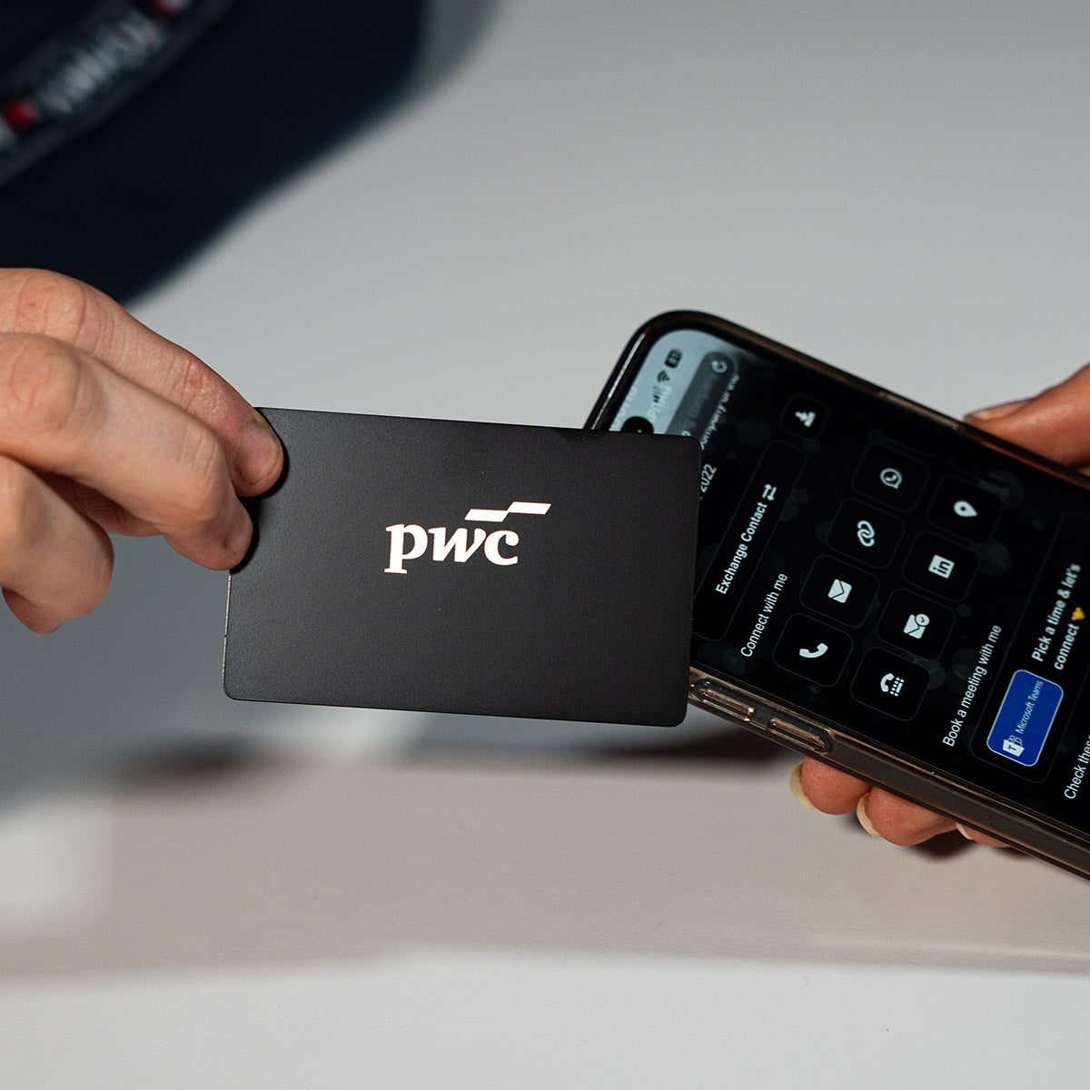 Close-up of a premium BLACK TAPiTAG metal digital business card wth PWC logo tapping a smartphone for instant contact sharing NFC