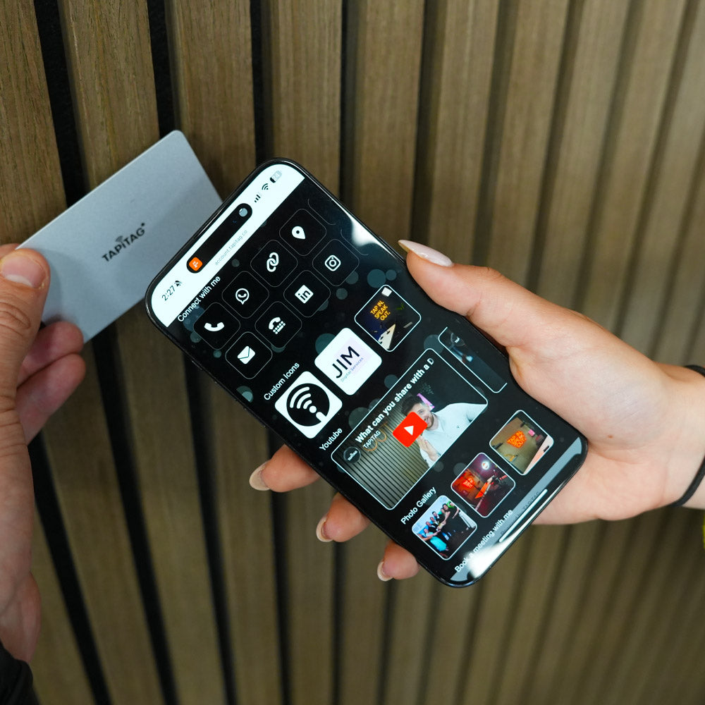 Person holding a smartphone with a black screen displaying app icons against a wooden background TAPiTAG Digital Business Card 