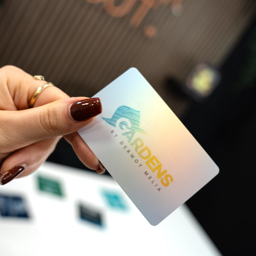 Hand holding a TAPiTAG Digital Business card card with 'Gardens by Dermot Melia' logo against a blurred background