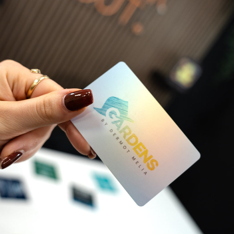 Hand holding a TAPiTAG Digital Business card card with 'Gardens by Dermot Melia' logo against a blurred background