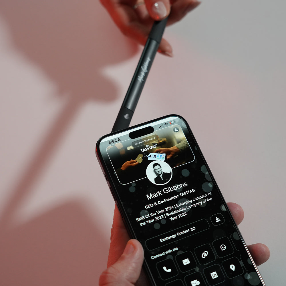 Person holding a smartphone with a contact card displayed on the screen. Phone is being held against a TAPiTAG Smart pen.