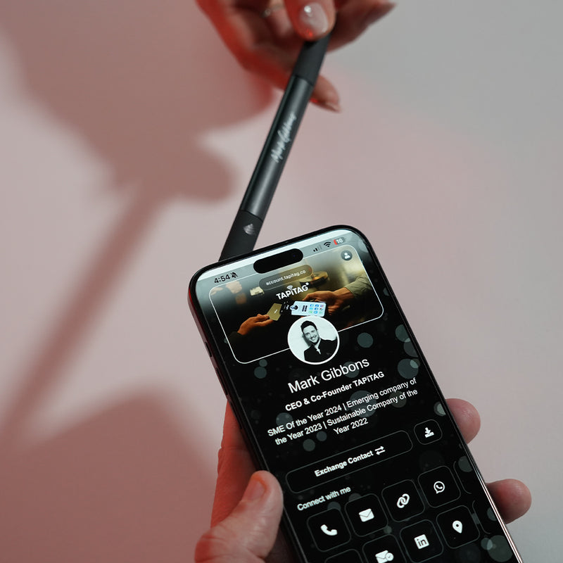 Person holding a smartphone with a contact card displayed on the screen. Phone is being held against a TAPiTAG Smart pen.