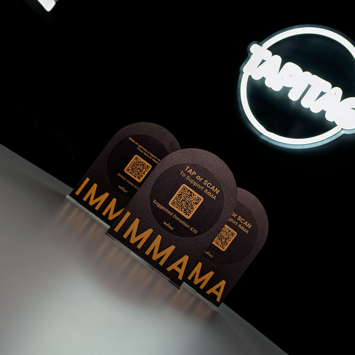 Two black cards with QR codes and 'IMMIMAMA' branding in front of a neon sign.