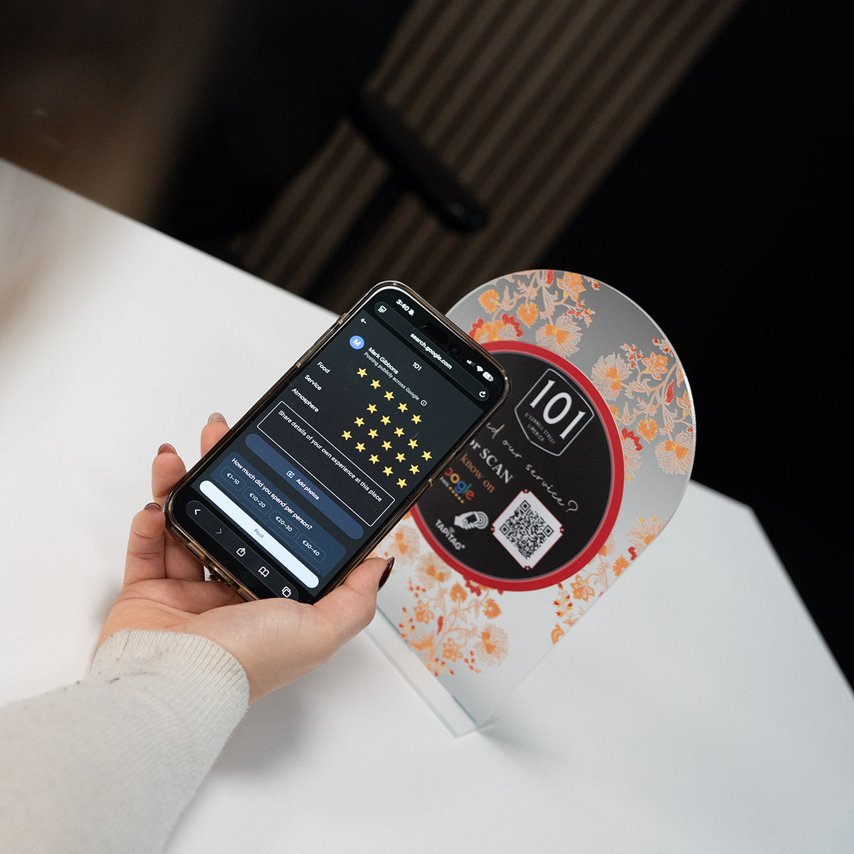 Hand holding a smartphone showing a Google review screen after tapping a TAPiTAG NFC stand on a restaurant counter, prompting customers to leave a 5-star review.