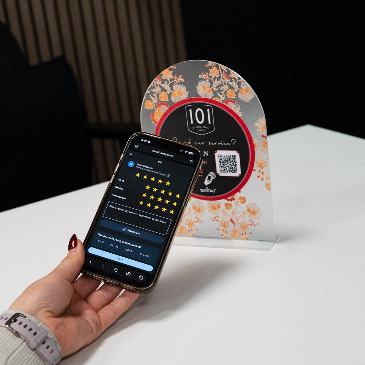 Hand holding a smartphone showing a Google review screen after tapping a TAPiTAG NFC stand on a restaurant counter, prompting customers to leave a 5-star review.