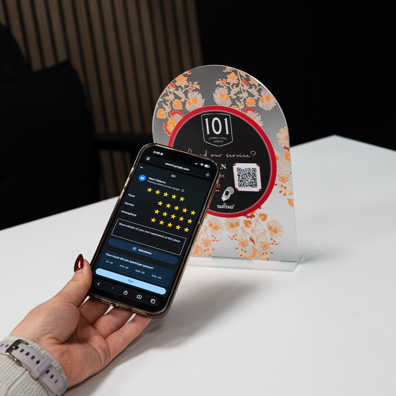 Hand holding a smartphone showing a Google review screen after tapping a TAPiTAG NFC stand on a restaurant counter, prompting customers to leave a 5-star review.