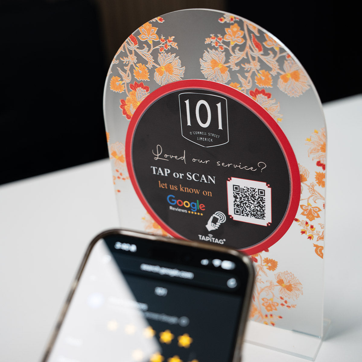 Hand holding a smartphone showing a Google review screen after tapping a TAPiTAG NFC stand on a restaurant counter, prompting customers to leave a 5-star review.
