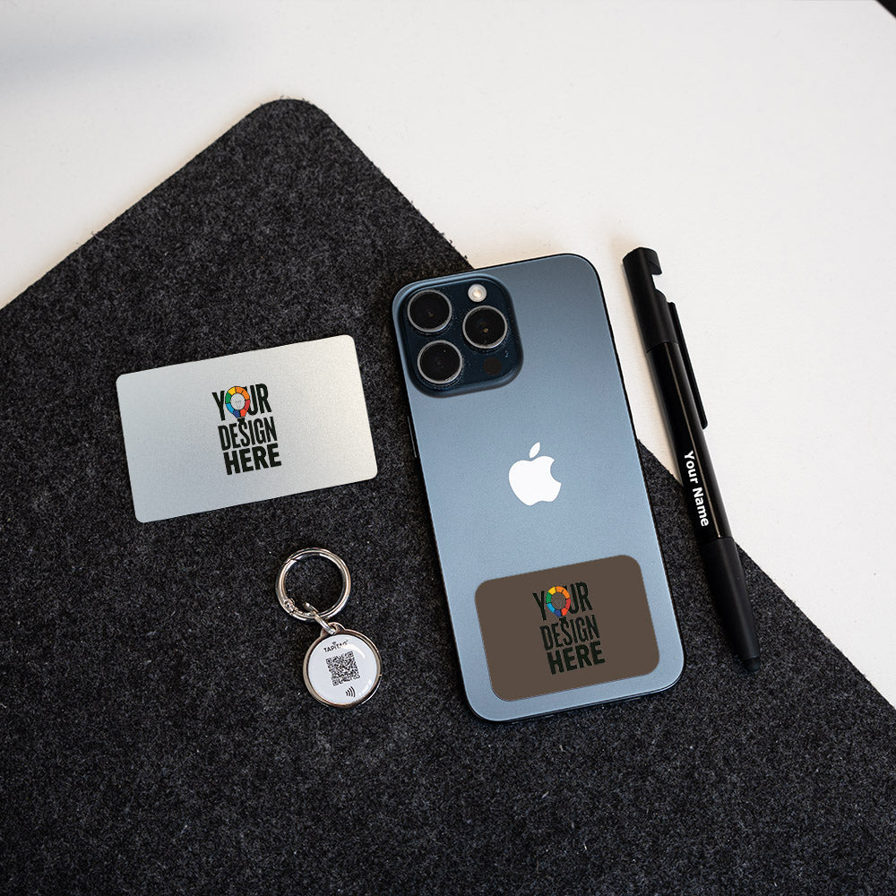 A dark slate blue iPhone case mockup displayed on a dark gray felt mat, alongside a TAPiTAG silver metal keychain, a TAPiTAG  white business card, and a TAPiTAG  black pen. The case features a custom brown rectangular patch with a colorful 'YOUR DESIGN HERE' graphic.
