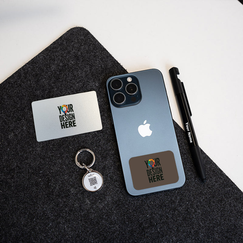 A dark slate blue iPhone case mockup displayed on a dark gray felt mat, alongside a TAPiTAG silver metal keychain, a TAPiTAG  white business card, and a TAPiTAG  black pen. The case features a custom brown rectangular patch with a colorful 'YOUR DESIGN HERE' graphic.