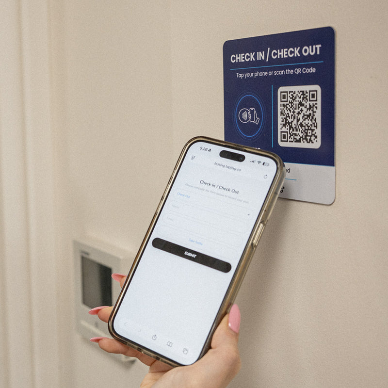 A hand holds a smartphone in front of a wall-mounted TAPiTAG QR code check-in/check-out kiosk. The kiosk has a dark blue top section with white text and a white lower section with a QR code, while the phone screen shows a check-in interface.
