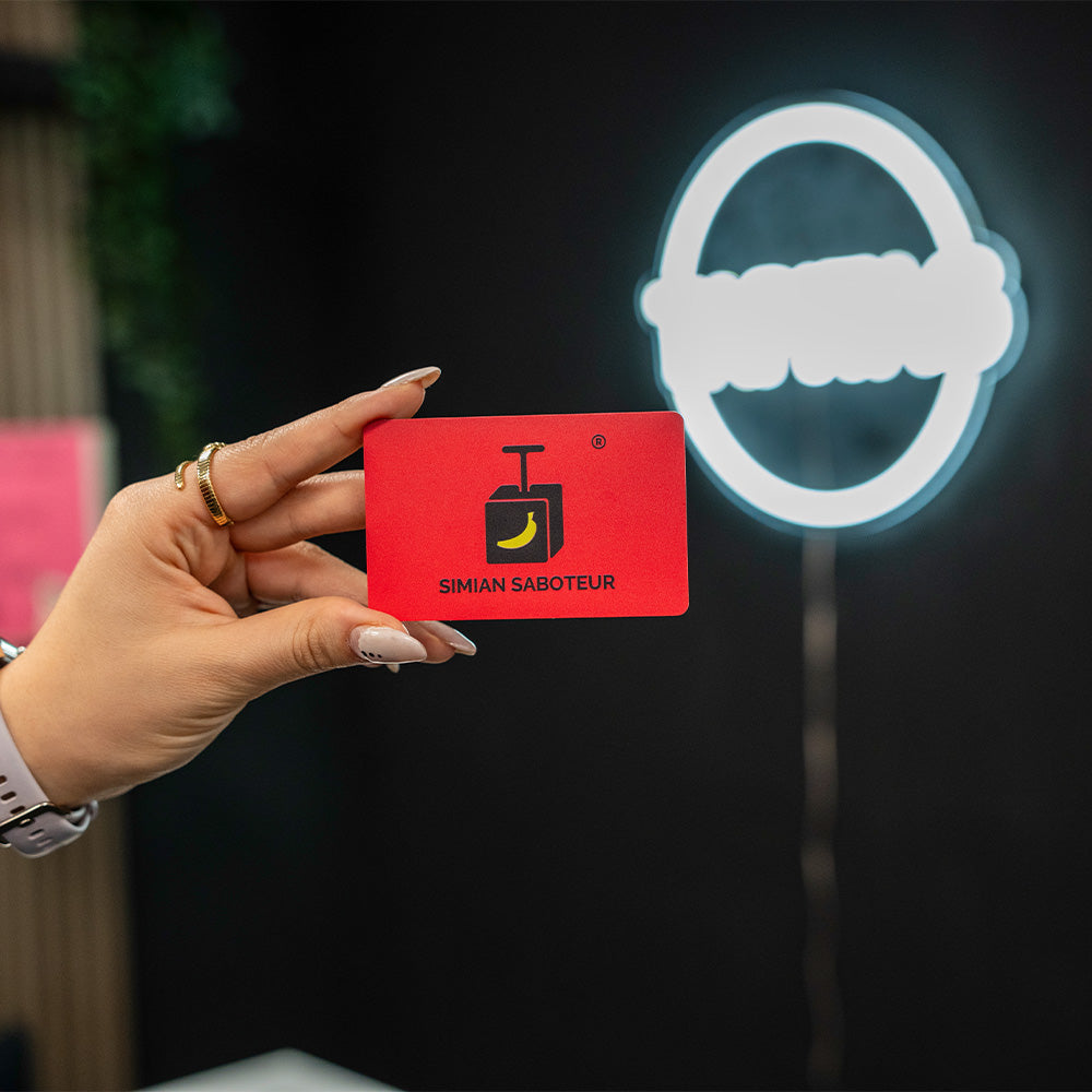 Hand holding a red TAPiTAG Digital Business Card with 'Simian Saboteur' logo in front of a dark background with a white symbol.