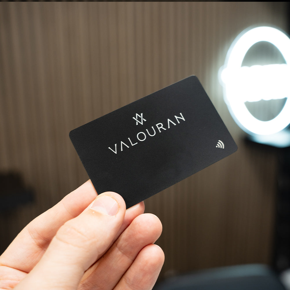 Hand holding a black TAPiTAG METAL DIGITAL BUSINESS card with 'VALOURAN' branding against a blurred background