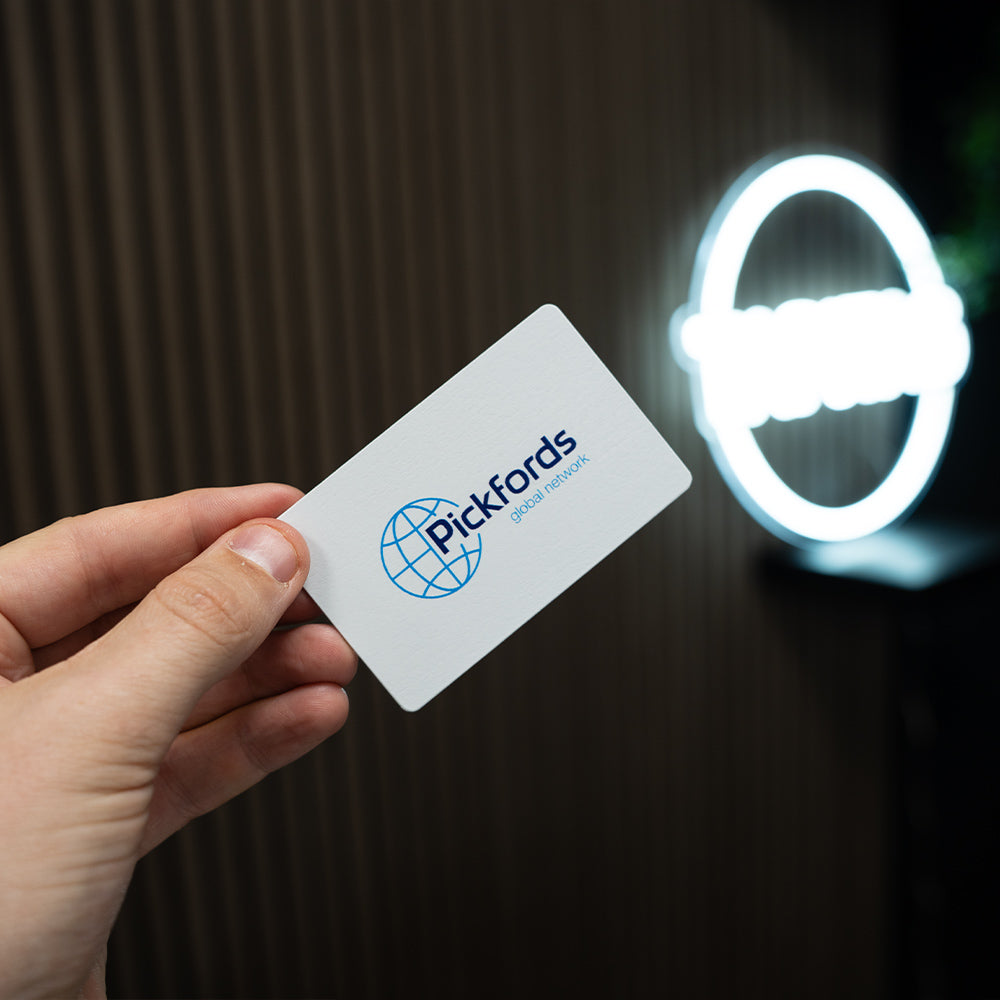 Hand holding a TAPiTAG Digital business card with 'Pickfords' logo in front of a neon light.
