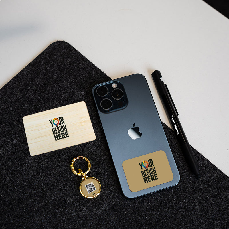 A dark gray smartphone case with a gold-toned rectangular patch reading 'YOUR DESIGN HERE' is shown on a phone, alongside a matching wooden NFC card, pen, and round keychain on a black felt surface against a white background.