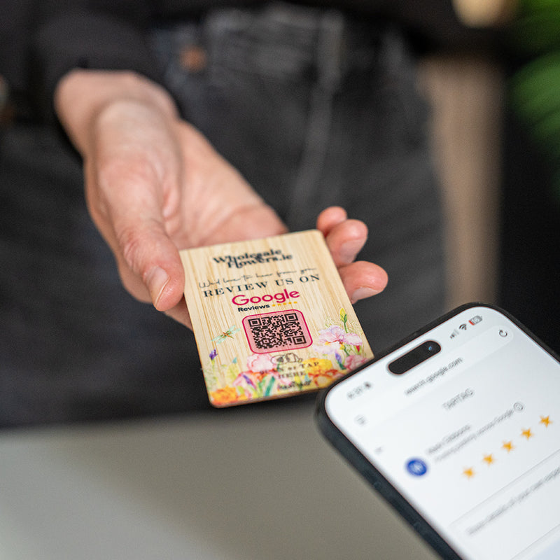 A hand holds a TAPITAG Bamboo wooden Google Review Plan card with colorful floral illustrations and a QR code, while a smartphone screen in the foreground shows the Google review page.