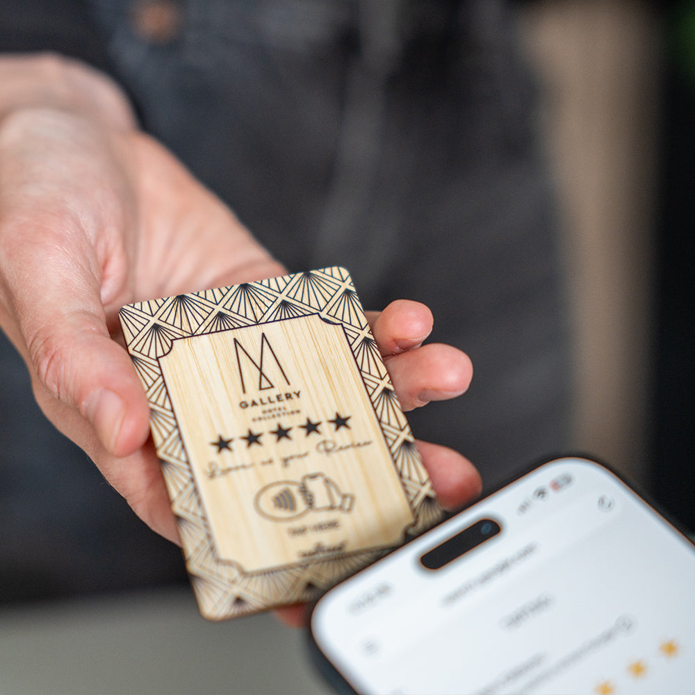 A hand holds a TAPITAG Bamboo wooden Google Review Plan card with colorful floral illustrations and a QR code, while a smartphone screen in the foreground shows the Google review page.