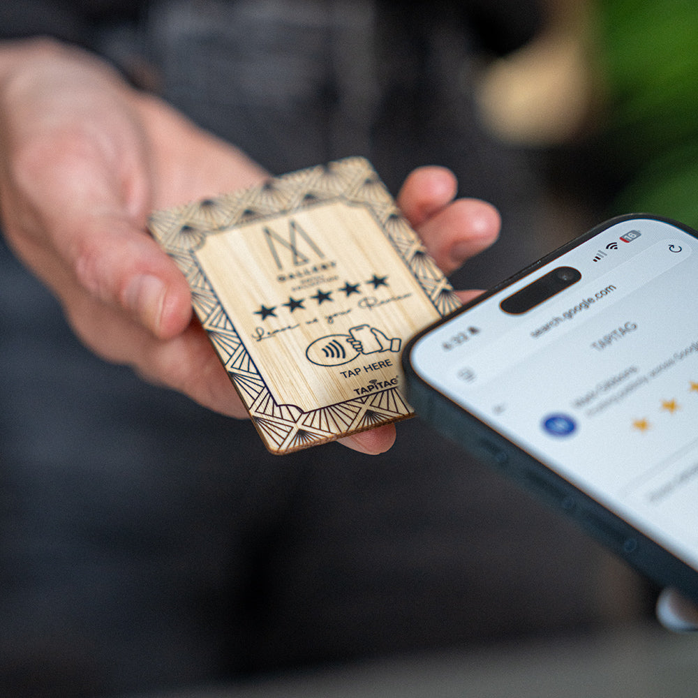 A hand holds a TAPITAG Bamboo wooden Google Review Plan card with colorful floral illustrations and a QR code, while a smartphone screen in the foreground shows the Google review page.