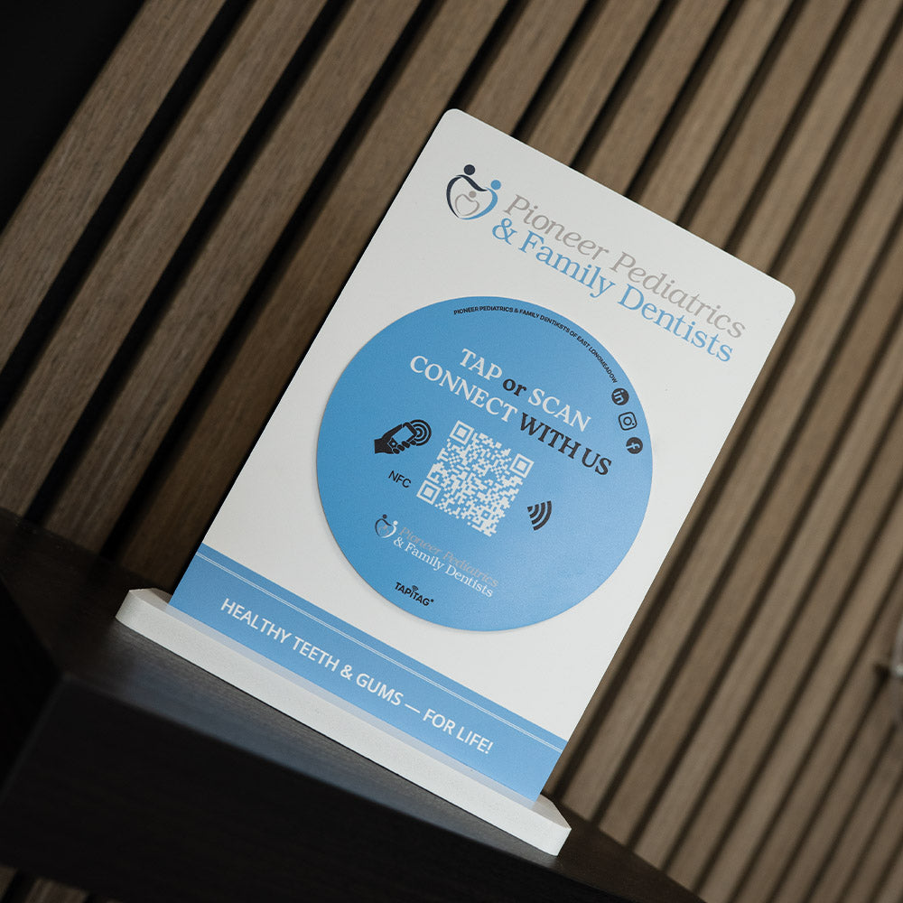 Card with Pioneer Pediatrics & Family Dentists branding and QR code on a wooden surface. Promotional sign for Pioneer Pediatrics & Family Dentists with a QR code on a blurred background. TAPiTAG NFC TAG Display stand
