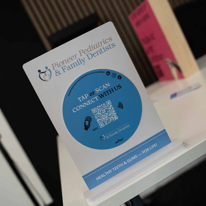 Promotional sign for Pioneer Pediatrics & Family Dentists with a QR code on a blurred background. TAPiTAG NFC TAG Display stand