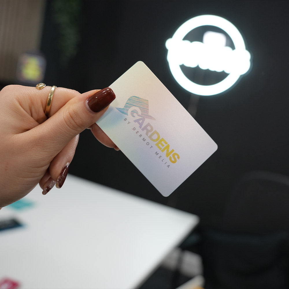 Hand holding a TAPiTAG Digital Business card card with 'Gardens' logo in front of a blurred background