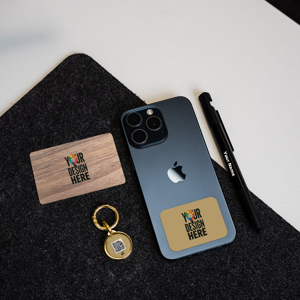 A dark gray smartphone case with a gold-toned rectangular patch reading 'YOUR DESIGN HERE' is shown on a phone, alongside a wooden card, black pen, and gold keychain on a black felt surface.