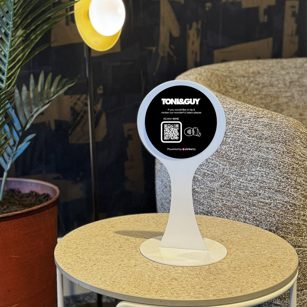 Round table with a white TAPiTAG Display stand holding a circular sign featuring 'TONI&GUY' branding in a room setting.