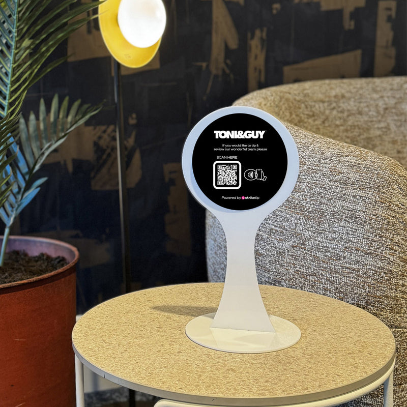 Round table with a white TAPiTAG Display stand holding a circular sign featuring 'TONI&GUY' branding in a room setting.