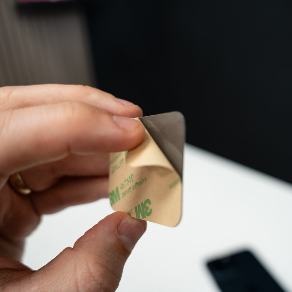 Hand holding a small adhesive strip with green text on a blurred . TAPiTAG NFC QR MINICARD NFC-Enabled Digital Business Cardbackground