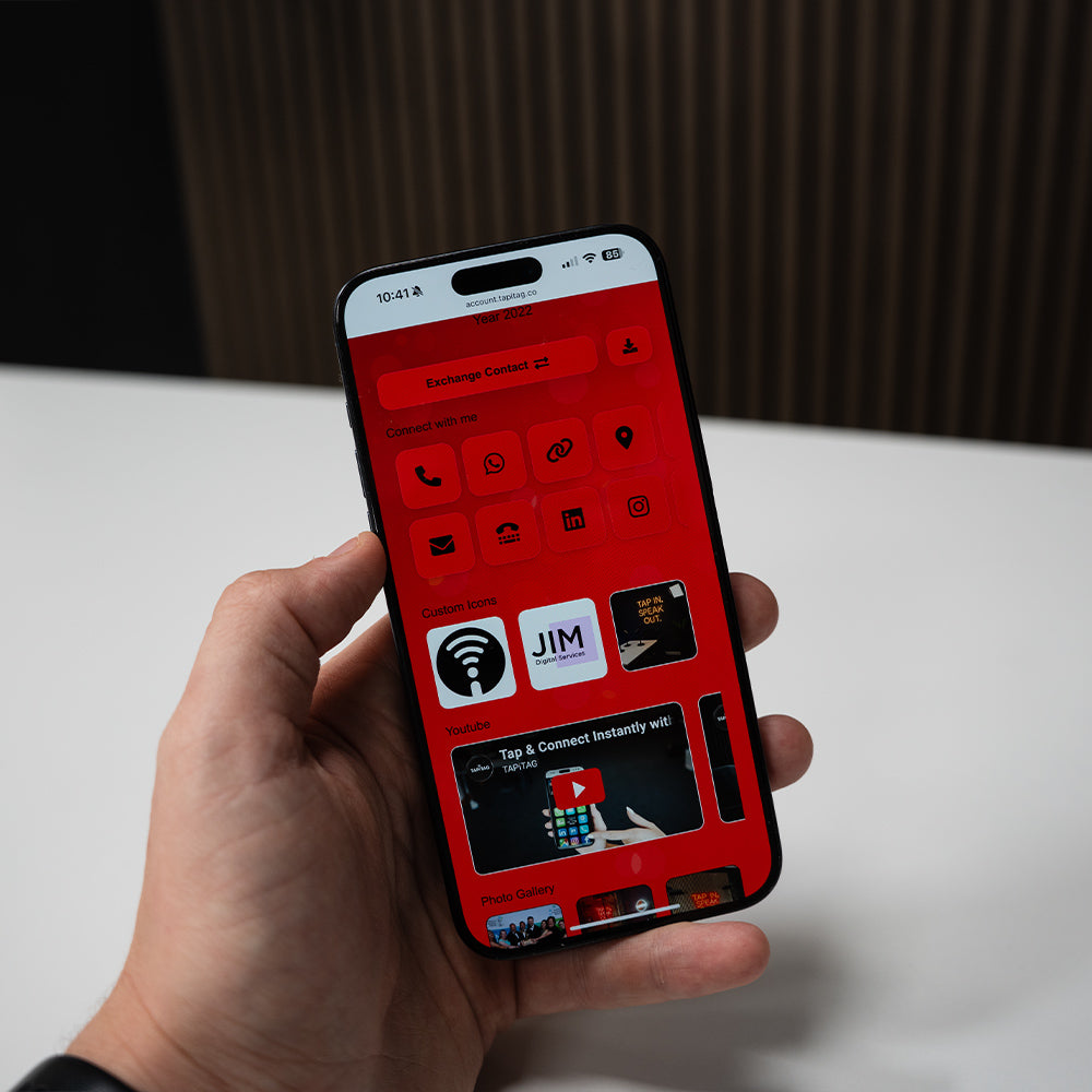 Person holding a smartphone with a red-themed app interface on a white surface. TAPiTAG Digital Business Card 