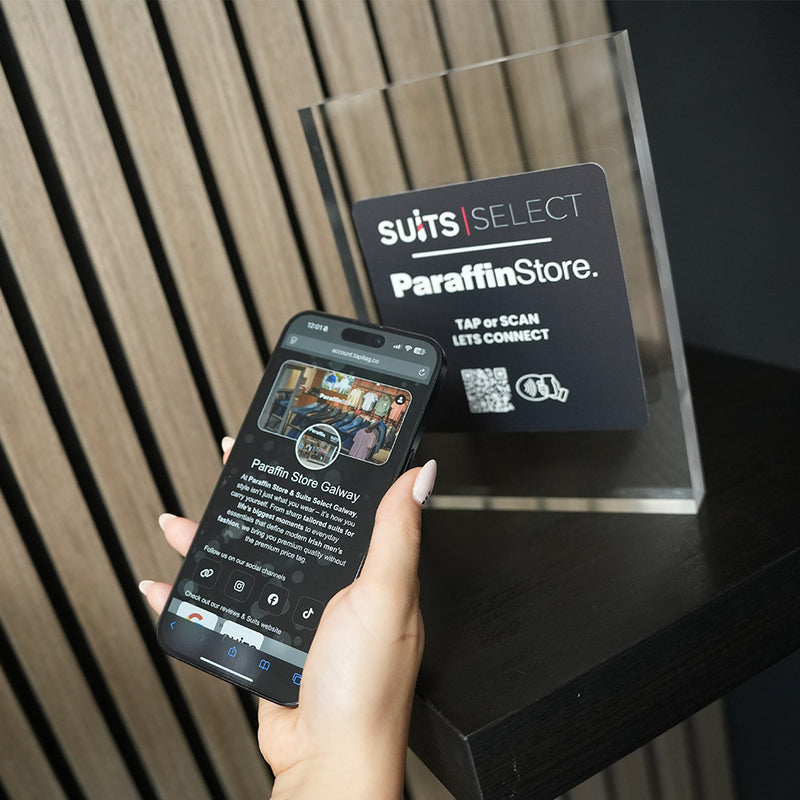 Hand holding a smartphone with a digital display next to a. 'SUITS SELECT Paraffin Store' sign Clear acrylic sign with 'SUITS SELECT Paraffin Store' branding on a reflective surface with nfc tag