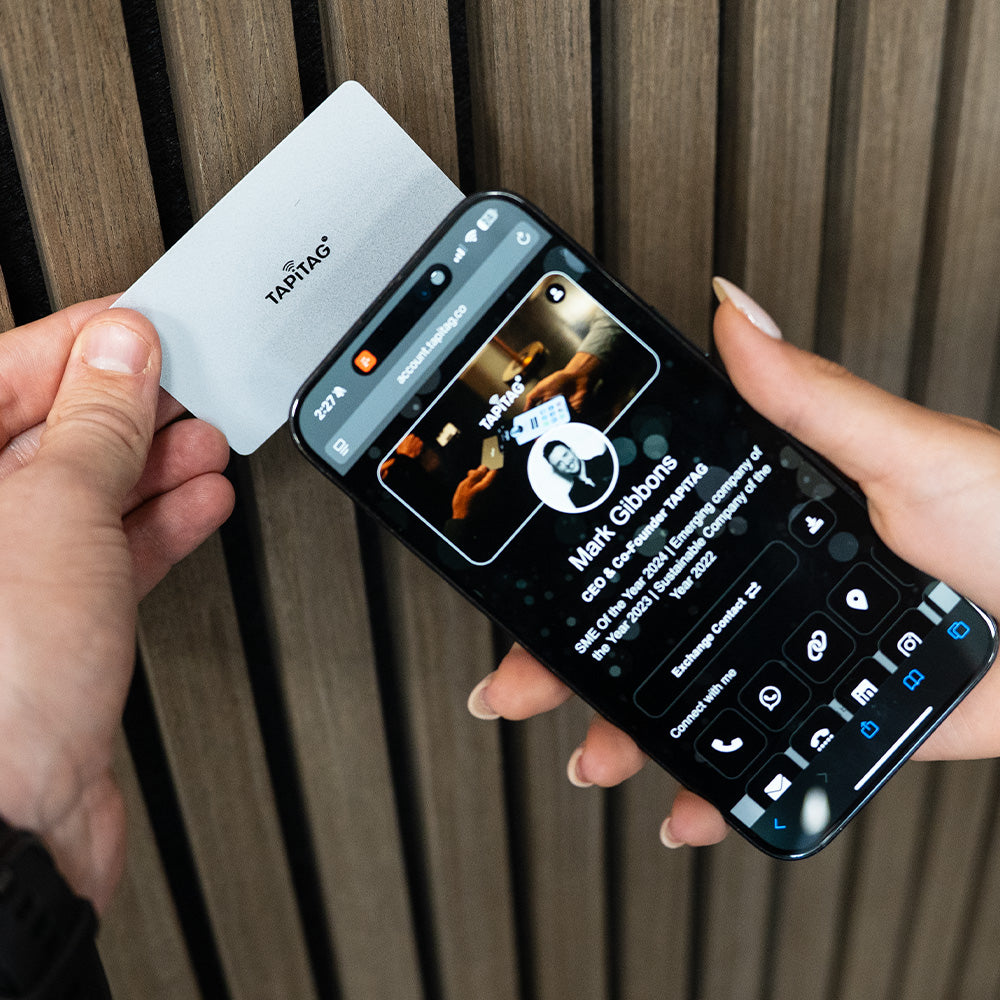 Person holding a smartphone with a call interface and a Tap Tag card against a wooden background TAPiTAG Digital Business Card 
