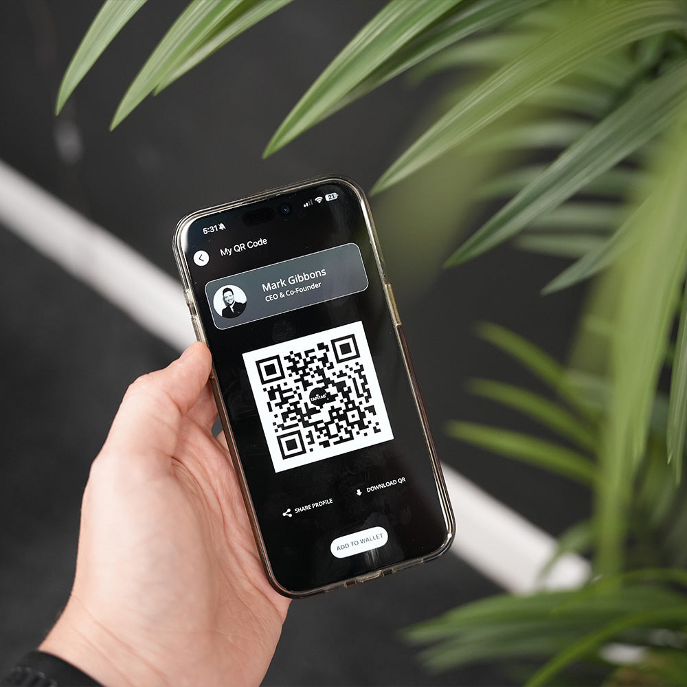 Hand holding a smartphone with a QR code displayed on the screen, against a blurred indoor plant background.