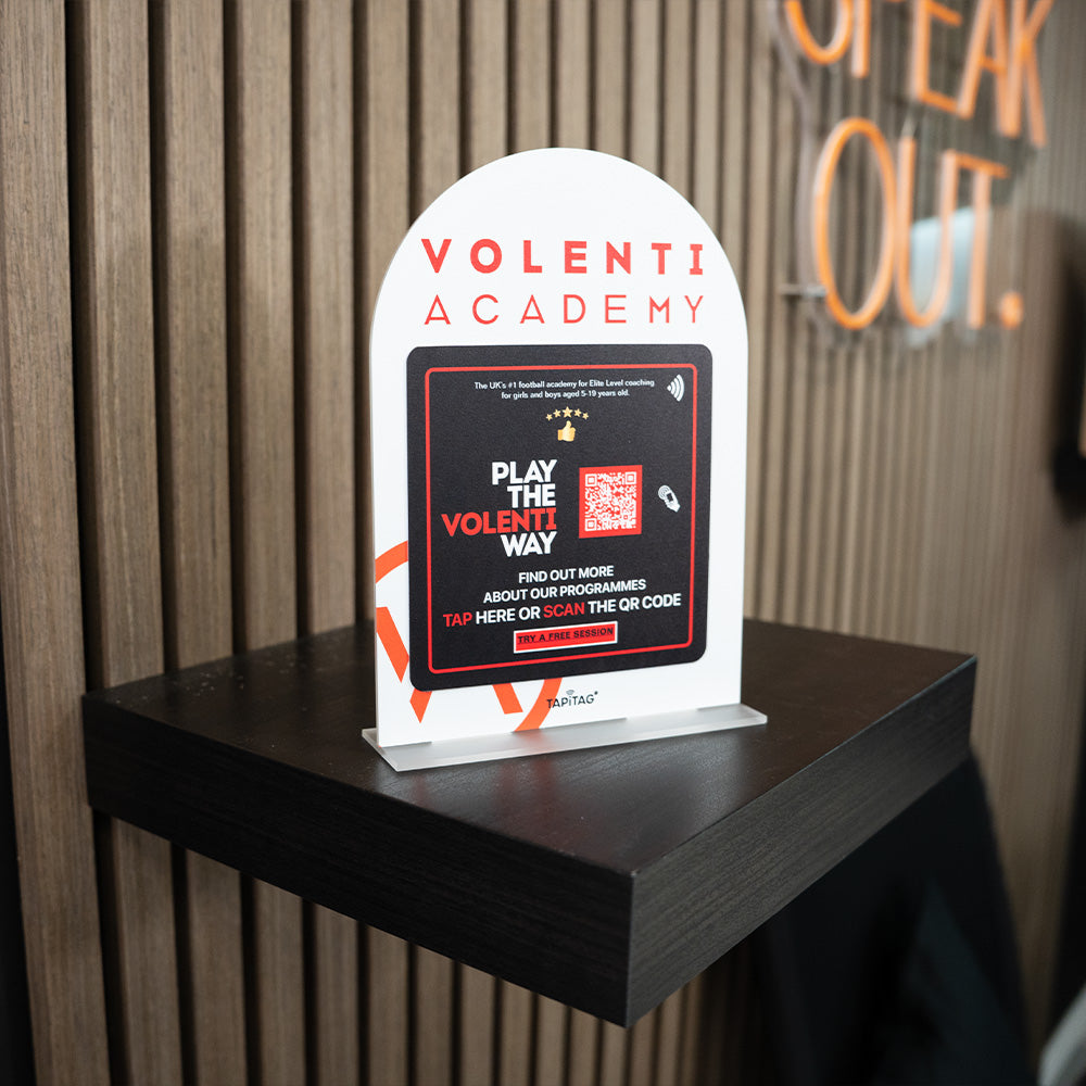 Promotional TAPiTAG Acrylic A5 ARCH display stand sign for Volenti Academy on a wooden shelf with a blurred background.