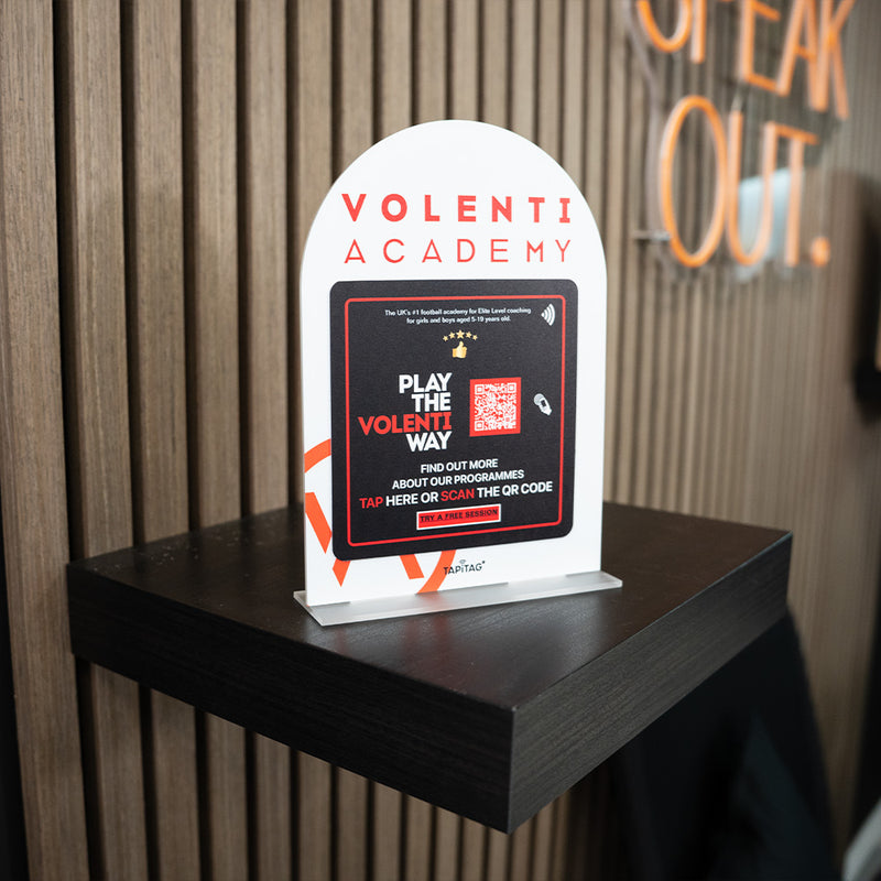 Promotional TAPiTAG Acrylic A5 ARCH display stand sign for Volenti Academy on a wooden shelf with a blurred background.