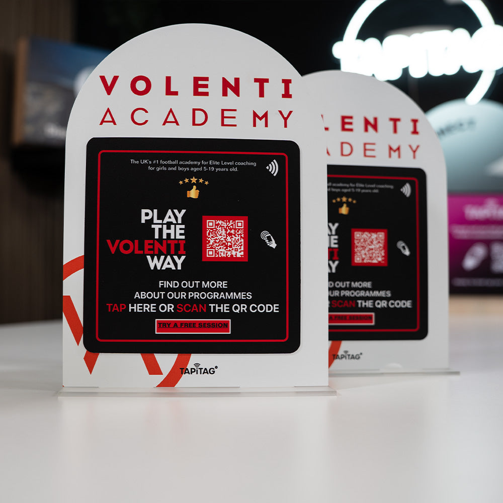 Two Volenti Academy promotional TAPiTAG Acrylic A5 ARCH display stand signs with QR code on a blurred background