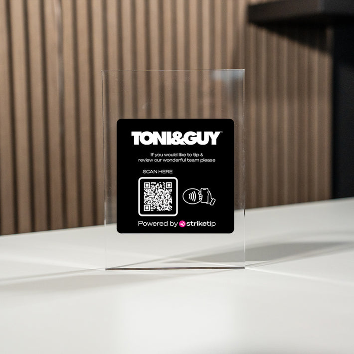 Toni&Guy tipping card with QR code on a clear TAPiTAG Display stand against a wooden wall.