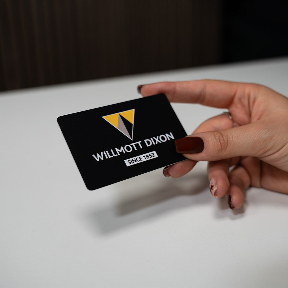 Hand holding a Willmott Dixon TAPiTAG Digital Business Card on a white surface with a dark background